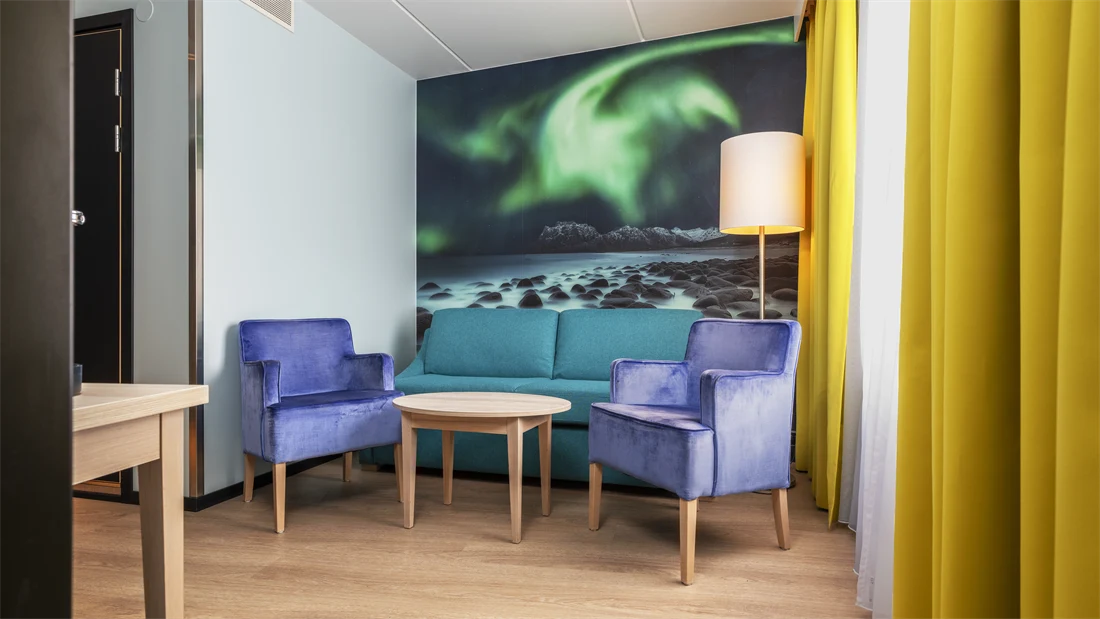 Junior suite med sittegruppe, lite rundt bord, gule gardiner og nordlys bilde på veggen i bakgrunnen