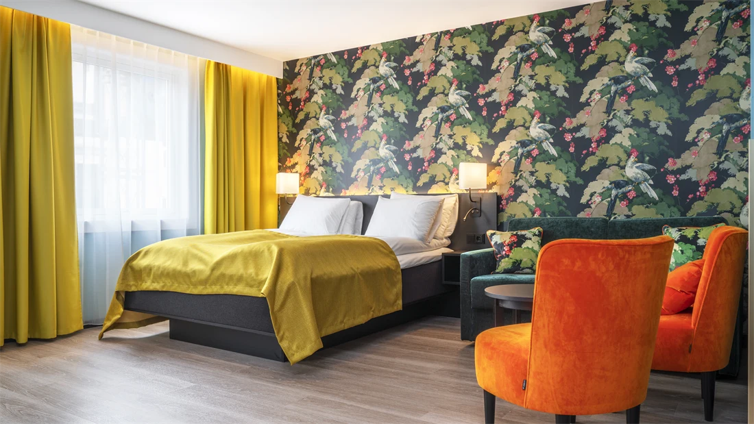Vindu med gule gardiner. Seng med gult sengeteppe. Fargerik tapet med fugler og blomster, grønn sofa, oransje stoler og mørkt bord i business rom på Thon Hotel Bristol Bergen