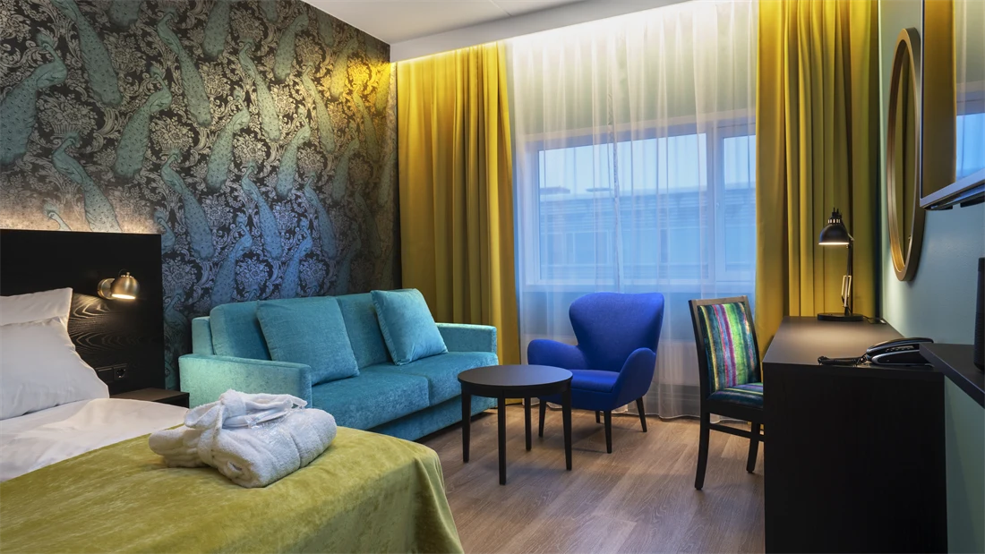 Parkettgulv, turkis sofa, to stoler, salongbord, skrivebord, dobbeltseng, vindu fra gulv til tak, gule gardiner, tapetvegg med mønster 