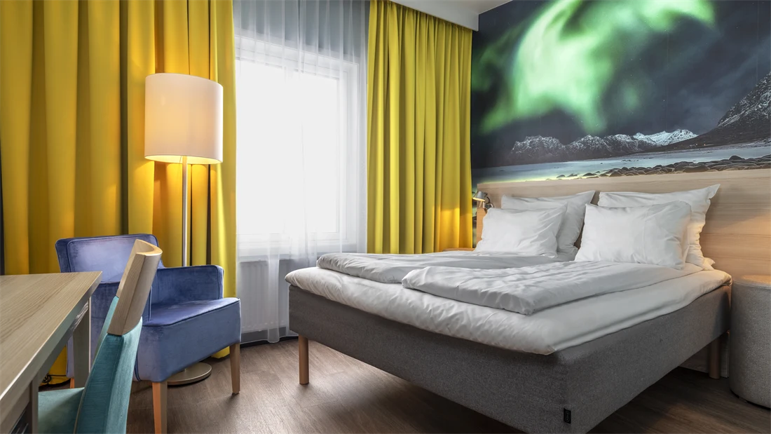 Dobbeltrom med dobbeltseng, trehvit sengegavl, stort nordlys bilde på veggen, gule gardiner, lilla lenestol, skrivebord med turkis stol.