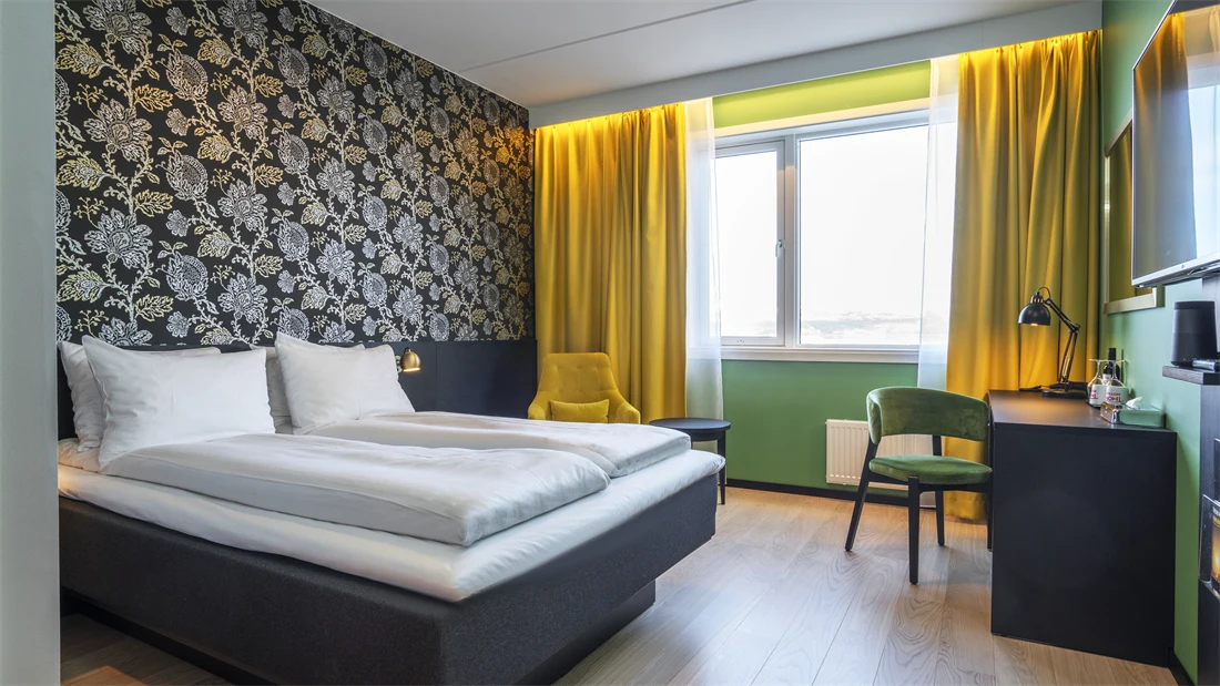 Dobbeltseng, mørk mønstrete tapet med blomster, gule gardiner, grønne vegger, skrivebord grønn stol og TV på dobbeltrom på Thon Hotel Kirkenes