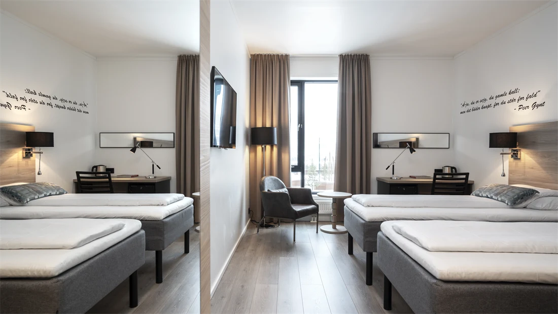 Helles Zweibettzimmer mit getrennten Betten, deckenhohen Fenstern, Schreibtisch, Sessel, Fernseher und Parkettboden. An der Wand hängt ein Zitat aus Peer Gynt.