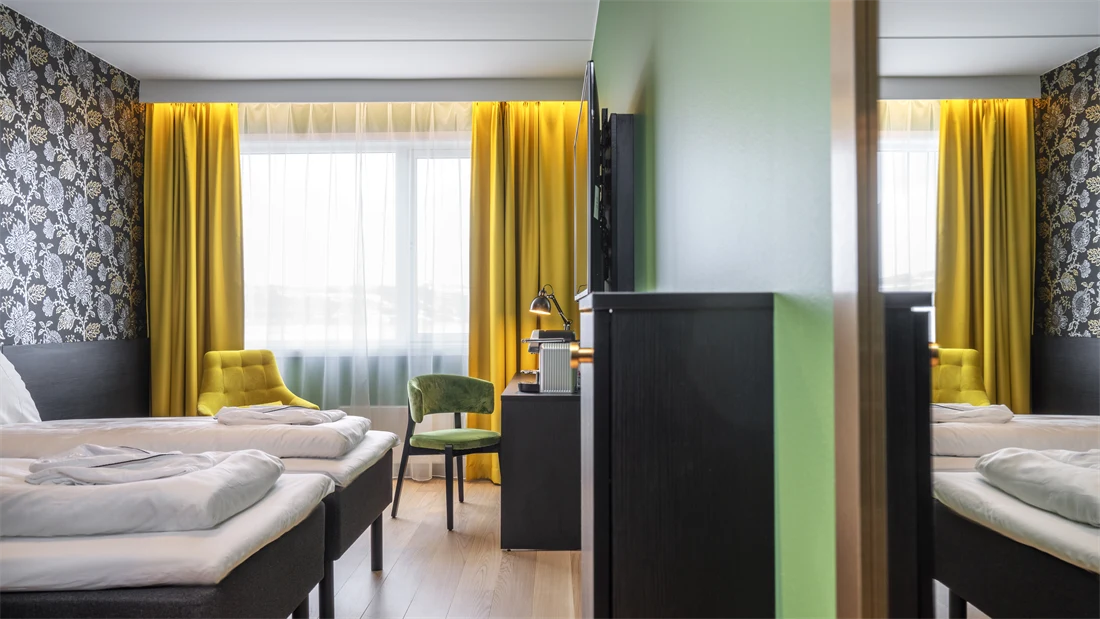 Separate senger, mørk mønstrete tapet med blomster, gule gardiner, gul stol, skrivebord med kaffemaskin og grønn stol i superior twin rom på Thon Hotel Kirkenes
