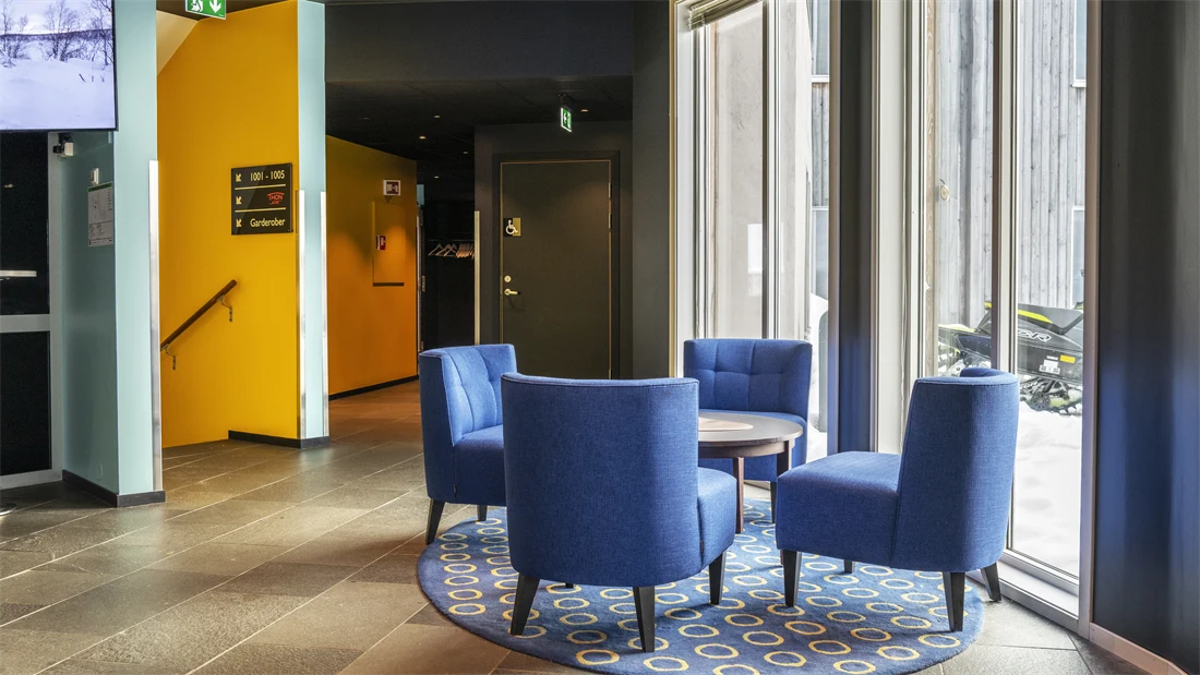 Lobbyområde med fire blå lenestoler rundt et rundt bord, rundt blått teppe under, og store vinduer ut.